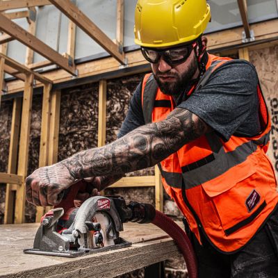 Construction worker with tattooed arm wearing safety gear operating circular saw on wooden beam inside building frame