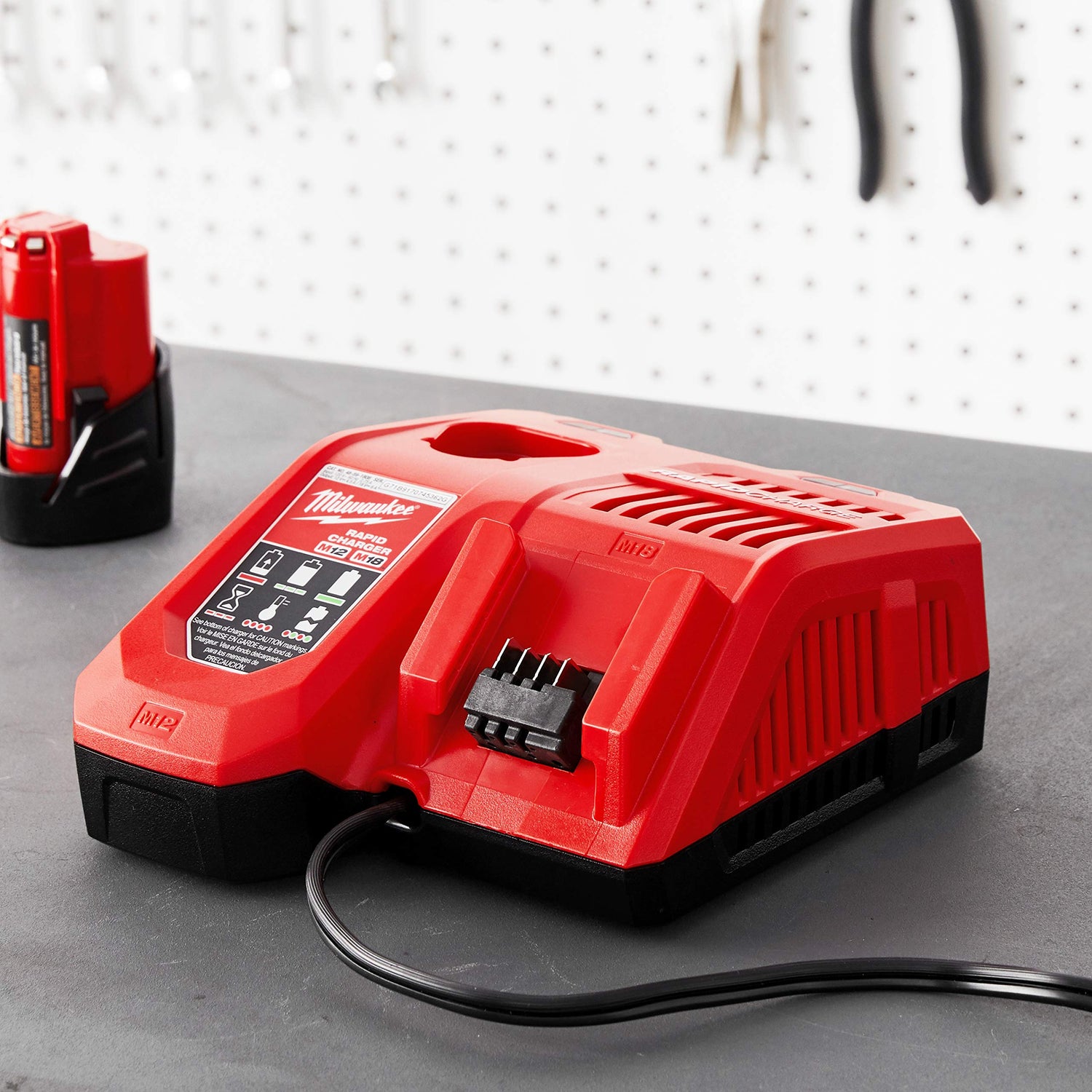 Red Milwaukee M18/M12 battery charger with cooling vents, status indicator panel, and power cord on gray workbench surface