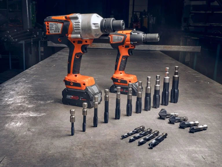 Two orange and black cordless impact wrenches with multiple drill and driver bits arranged on a metal work surface in workshop.