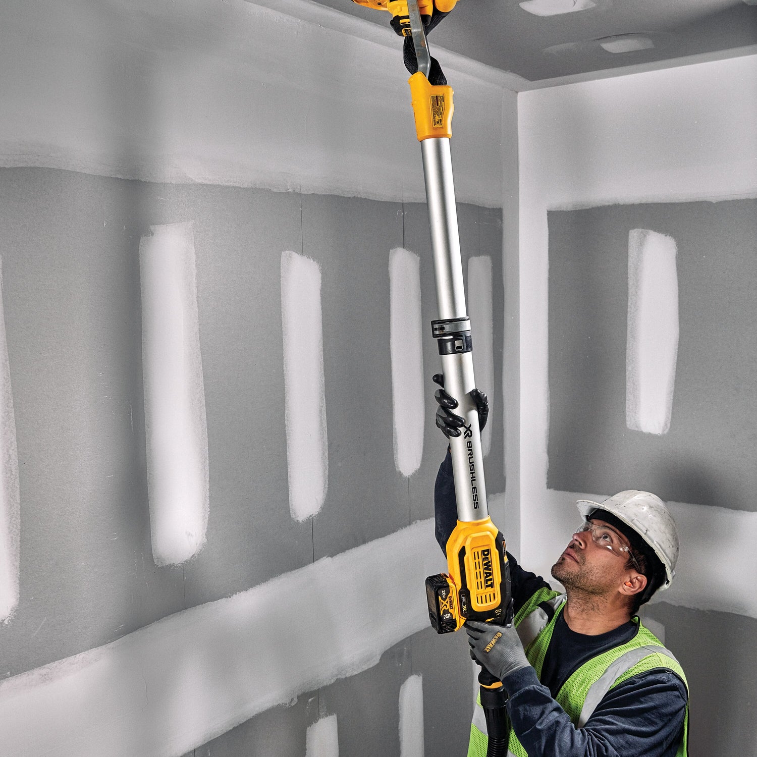 Cordless drywall sander with brushless motor and dust extractor attachment used by worker in safety gear on ceiling drywall panels