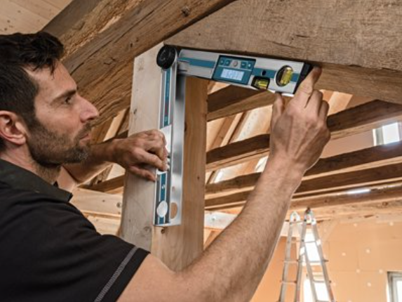 Man using Bosch digital inclinometer GAM 220 MF with illuminated pivoting screen to measure wooden beam angle in construction site